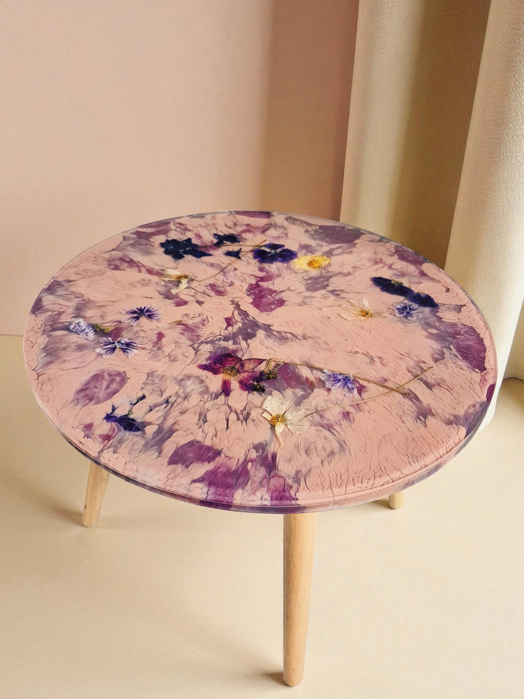 Rundtisch „Violet Veil“ aus Epoxidharz mit eingebetteten Blüten in Rosa- und Violetttönen, aufgestellt in neutraler Umgebung mit beigem Hintergrund.