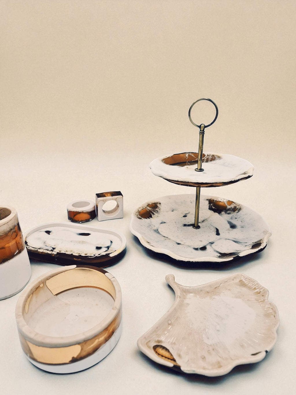 Kollektion „Melted Sun“ bestehend aus handgegossenen Wohnaccessoires aus Jesmonite und Epoxidharz in Weiß- und Goldtönen. Im Bild zu sehen sind Etagere, Schale, Serviettenringe, Tablett, Ginkgoblatt-Schale und weitere Dekoelemente auf hellem Hintergrund – harmonisch arrangiert im warmen Licht.