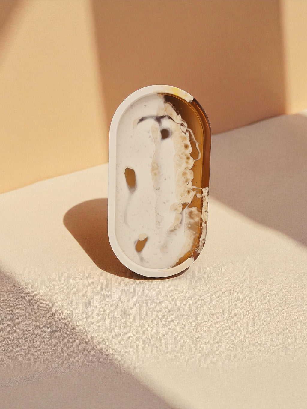 Ovales Tablett „Melted Sun“ aus Jesmonite und Epoxidharz mit marmorierten Farbverläufen in Weiß, Creme und Gold, auf hellem Stoff fotografiert.